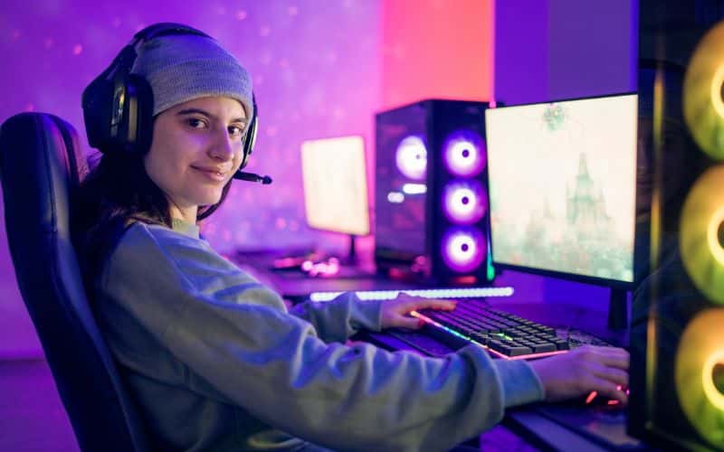 Fille devant un bureau avec un PC fixe gamers, elle porte un casque audio avec micro, elle a des accessoires gamers avec de la lumière néon d'ambiance sur la tour, la souris, le clavier et même le tapis de la souris.