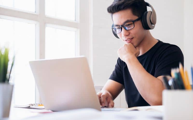 Homme portant des lunettes, un casque audio, devant un ordinateur portable, avec des papiers à côté de lui.