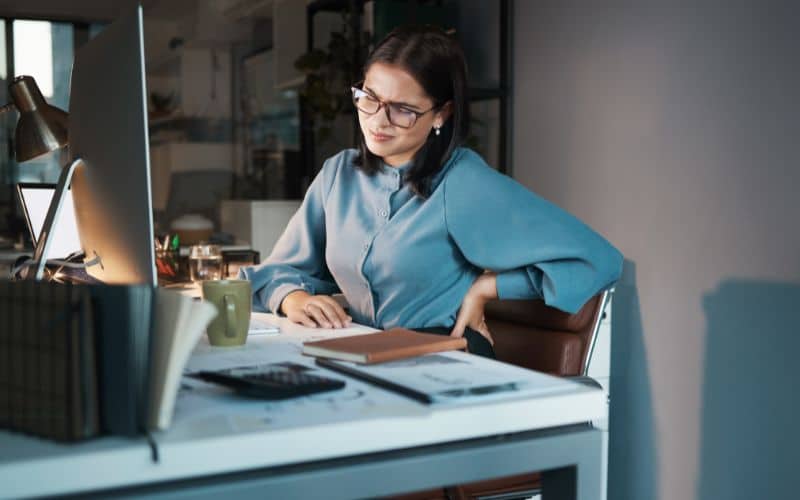 Femme assise à un bureau devant un ordinateur avec des papiers et carnet de notes, elle se tient le bas du dos et fait la grimasse.