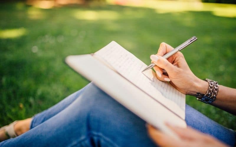 Personne dehors assise dans l'herbe, avec un cahier ouvert qui écrit avec un stylo ergonomique.