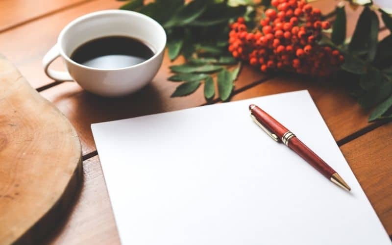 Feuille blanche posée sur une table en bois, avec un stylo ergonomique posé dessus, à côté d'une tasse de café et des fleurs.