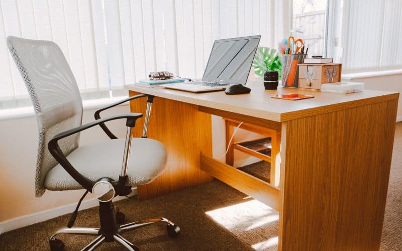 Bureau en bois avec un ordinateur portable, une chaise ergonomique devant, le tout installé près d'une grande fenêtre avec des voilages.